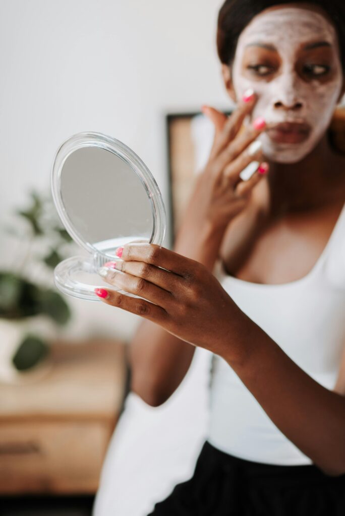 pexels-photo-9832152-9832152 A woman applies a white face mask while holding a handheld mirror in a cozy bedroom setting.